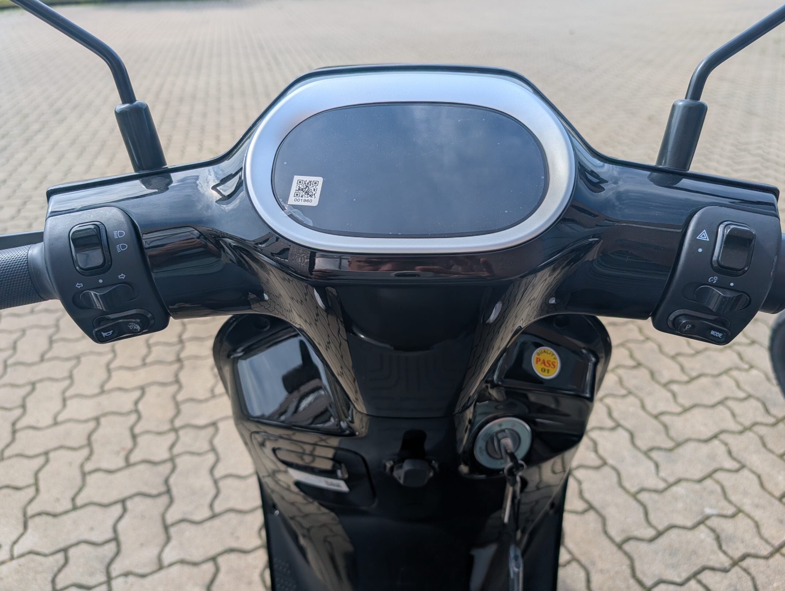 Nahaufnahme des Lenkers und des Armaturenbretts des schwarzen Yadea Owin U 2800W Retro-Elektrorollers (45 km/h) mit Bedientasten, einem Schlüssel im Zündschloss und einem auf dem Display sichtbaren QR-Code. Im Hintergrund ist eine gepflasterte Fläche zu sehen.