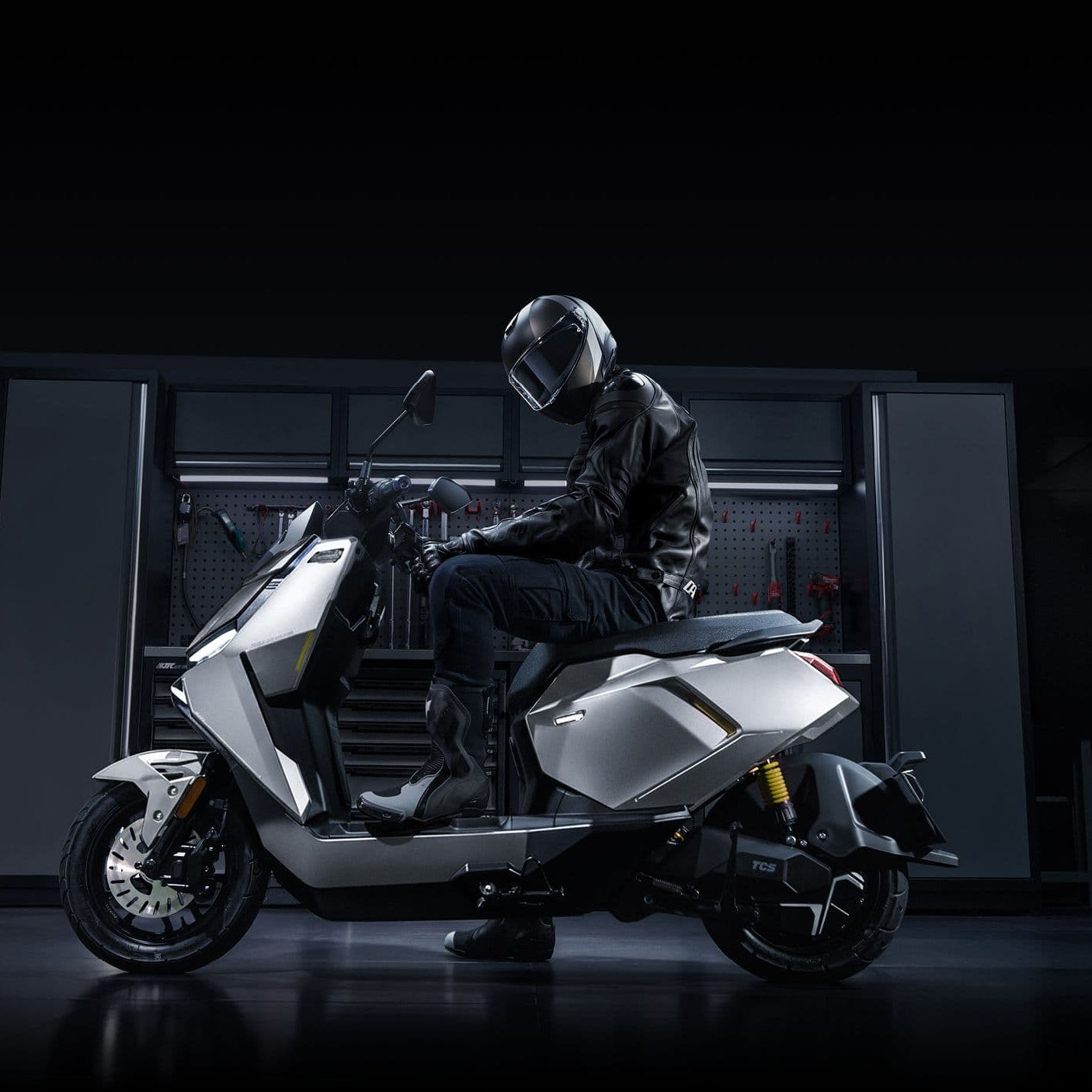 Eine Person mit Helm und schwarzer Jacke sitzt auf dem NIU FQiX 300 Elektroroller 80 km/h mit 3600W Motor und Dual-Akku von NIU in einer schummrig beleuchteten Garage mit Werkzeugschränken im Hintergrund.