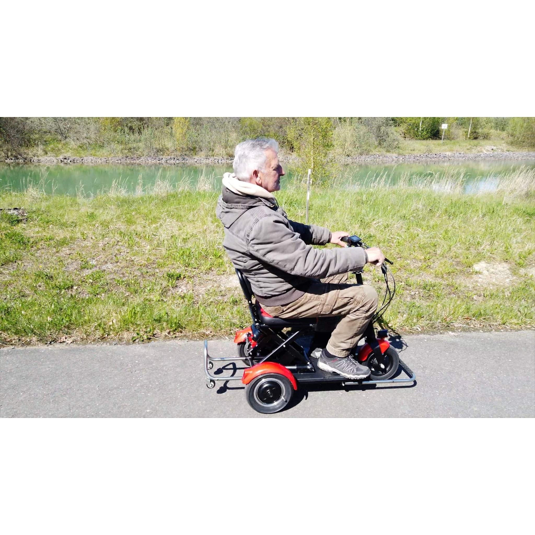 Ein älterer Mann in brauner Jacke und khakifarbener Hose fährt mit einem rot-schwarzen SONS OF ELECTRICITY Mobot Krankenfahrstuhl/Seniorenmobil 15 km/h - faltbar, leicht & mit Straßenzulassung über einen gepflasterten Weg durch Gras mit Wasser im Hintergrund.