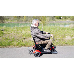 Ein älterer Mann in einer braunen Jacke fährt mit dem SONS OF ELECTRICITY Mobot Krankenfahrstuhl / Seniorenmobil 15 km/h - faltbar, leicht & mit Straßenzulassung auf einem gepflasterten Weg durch grünes Gras und einen Fluss und genießt den sonnigen Tag.