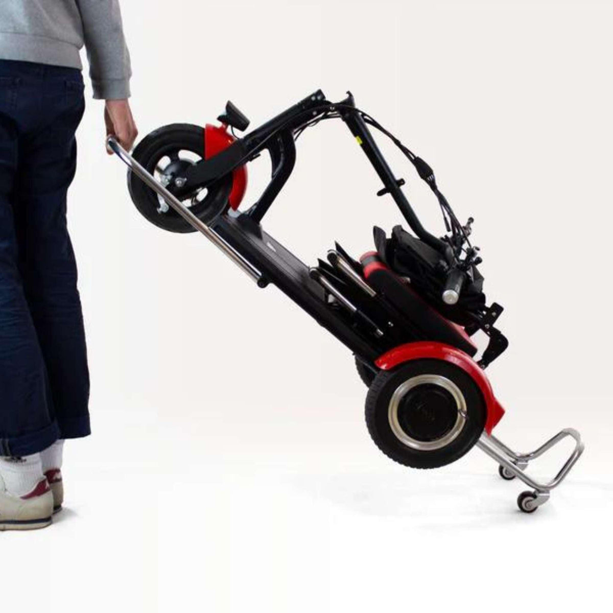 Eine Person in blauer Hose und weißen Schuhen zieht ein zusammengeklapptes SONS OF ELECTRICITY Seniorenmobil / Mobilitätshilfe Mobot PRO 10,5 Ah 3Rad - zusammenklappbar auf zwei kleinen Rädern an seinem Griff vor einem schlichten weißen Hintergrund.