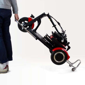 Eine Person in blauer Hose und weißen Schuhen zieht ein zusammengeklapptes SONS OF ELECTRICITY Seniorenmobil / Mobilitätshilfe Mobot PRO 10,5 Ah 3Rad - zusammenklappbar auf zwei kleinen Rädern an seinem Griff vor einem schlichten weißen Hintergrund.