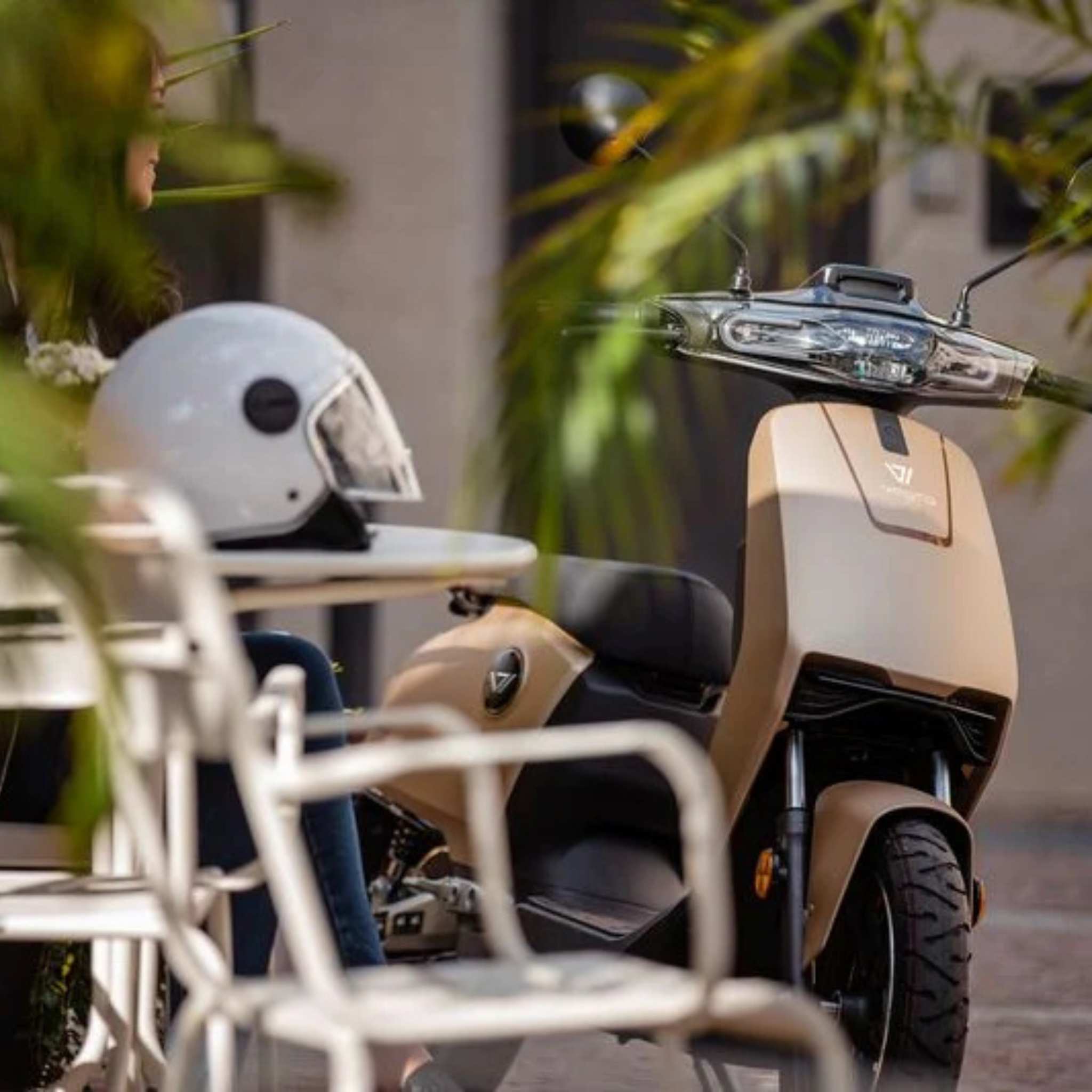 Ein beiger Super Soco CUx Pro Elektroroller mit Bosch-Power (45 km/h E-Roller) steht neben einem Tisch mit weißem Helm. Eine Person sitzt in der Nähe, teils von grünen Blättern verdeckt. Die Szene draußen betont das schlanke Design des E-Scooters.