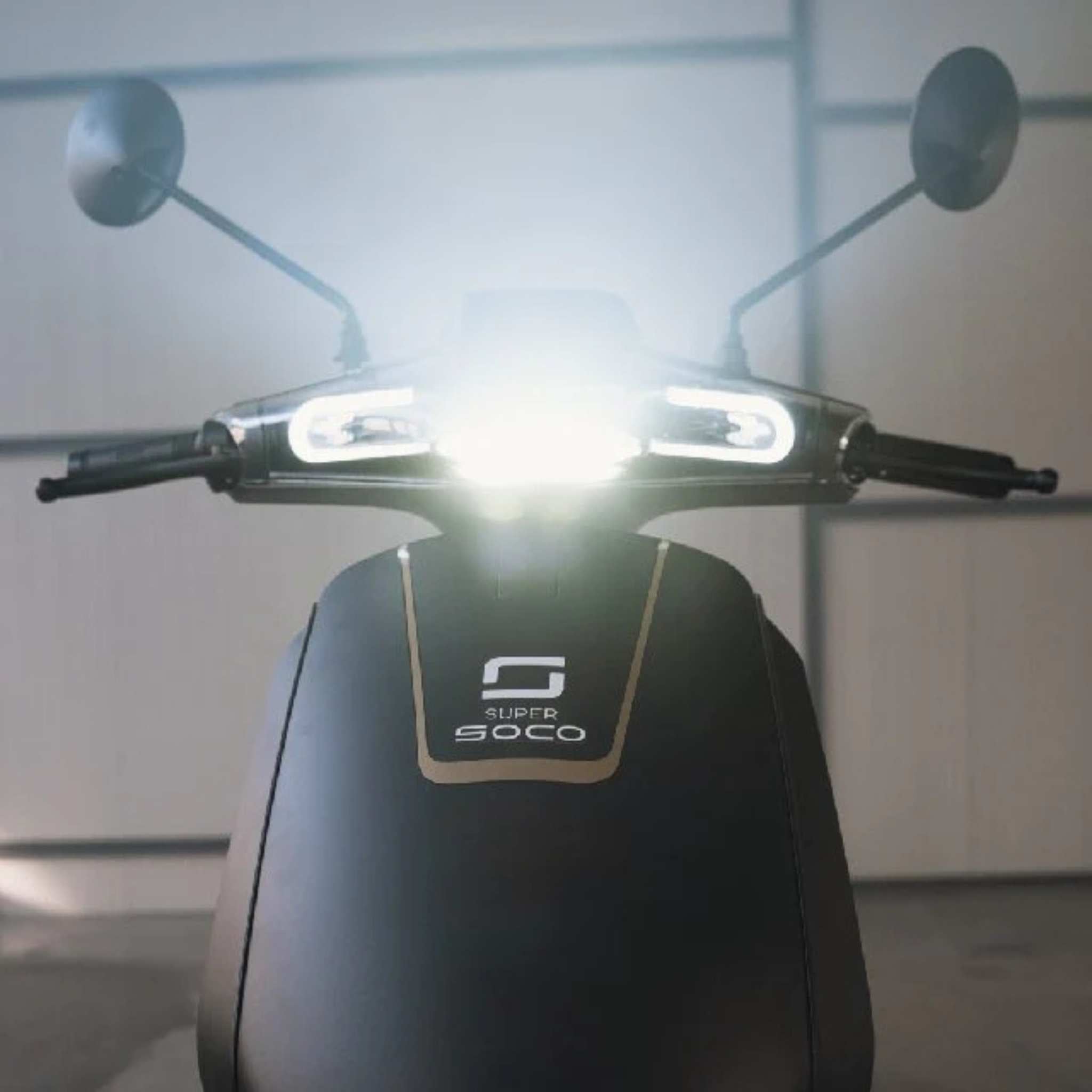 Eine Nahaufnahme des Super Soco CUx Pro Elektrorollers mit eingeschalteten Scheinwerfern und Lenkerleuchten, geparkt in einem Innenraum vor einer Lichtwand, verdeutlicht die Bosch-Power Technologie in diesem 45 km/h leichten E-Roller von Super Soco.
