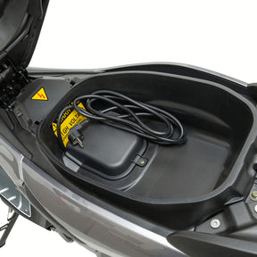 Offenes Fach unter dem Sitz eines Trinity Jupiter GT 300 Elektrorollers, in dem ein aufgerolltes Stromkabel und ein gelbes Hochspannungs-Warnschild zu sehen sind.