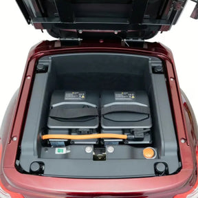 Eine Nahaufnahme des vorderen Kofferraums („Frunk“) eines TRINITY Atlas Premium 1800 zeigt zwei Akkupacks und Komponenten aus der Komfort-Serie, wobei die Motorhaube geöffnet ist und die umgebende Karosserie in Metallic-Rot lackiert ist.