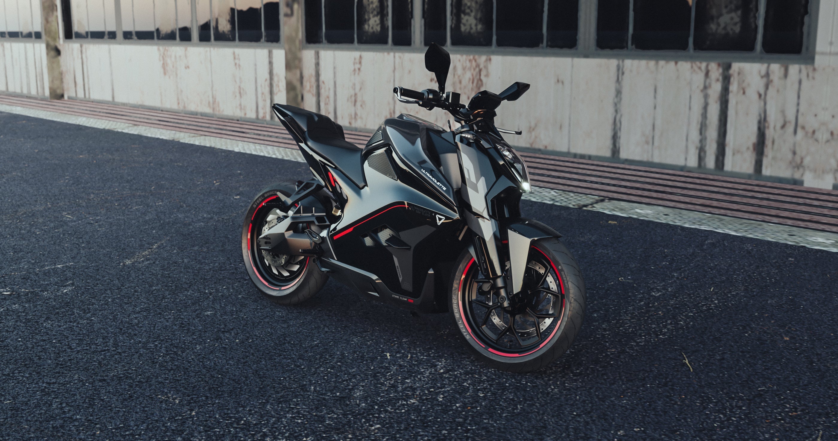 Ein auffälliges Ultraviolette F77 Superstreet Recon - Sportliches Elektromotorrad steht auf dunklem Asphalt vor einem verwitterten Gebäude, seine schwarz-silberne Karosserie mit roten Akzenten zieht die Blicke auf sich - bereit für die Straßen der Stadt.