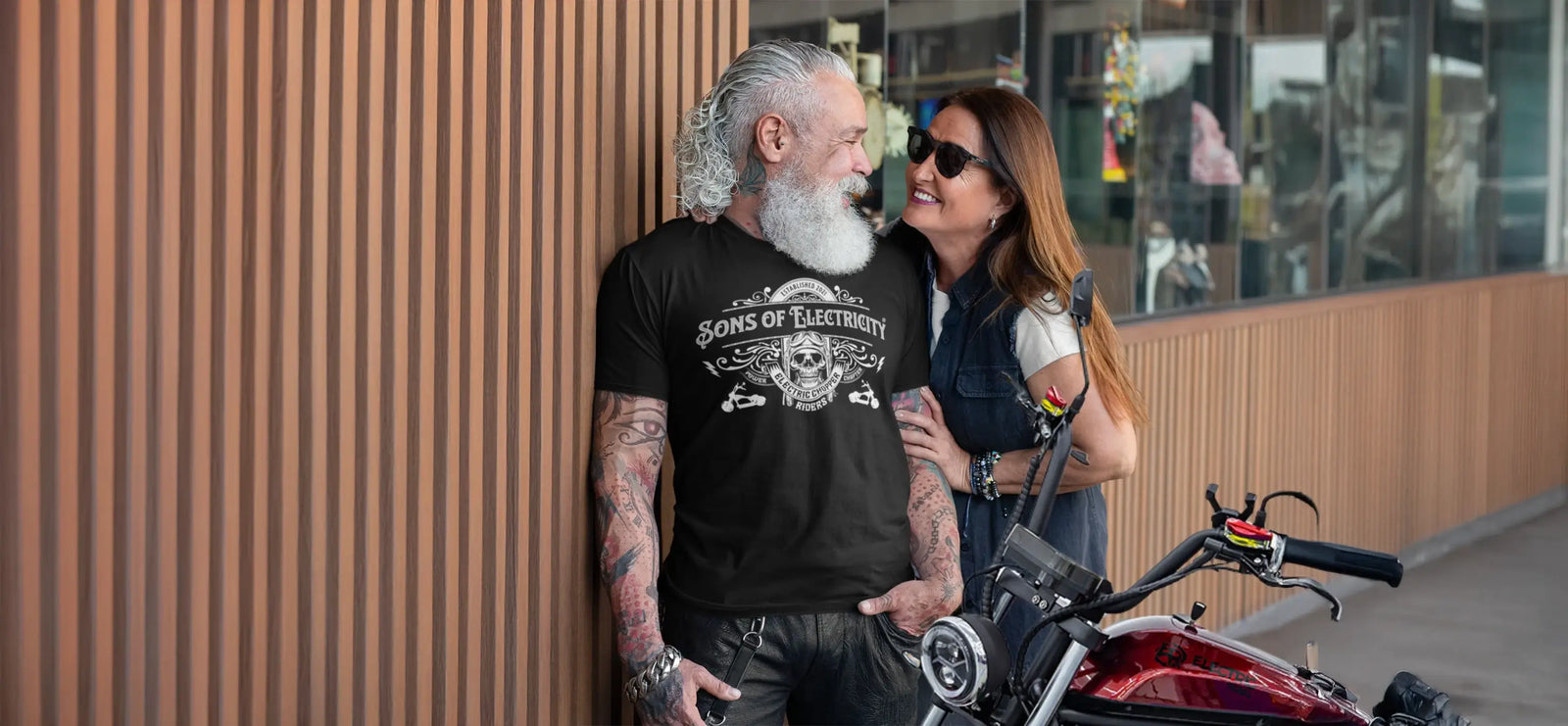 Älteres Paar lächelt sich an, Mann im schwarzen T-Shirt mit Aufdruck Sons of Electricity neben einem E-Chopper