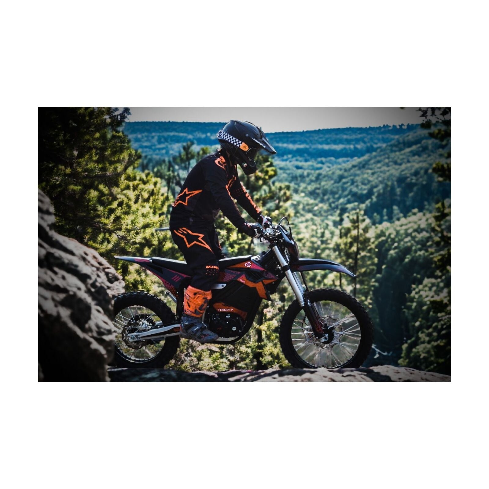 Ein Fahrer in schwarzer und oranger Kleidung fährt mit dem Trinity Panthera II Enduro - Elektromotorrad mit 1143 Nm Drehmoment und Schaltgetriebe durch einen felsigen Waldweg, im Hintergrund erheben sich grüne Hügel.