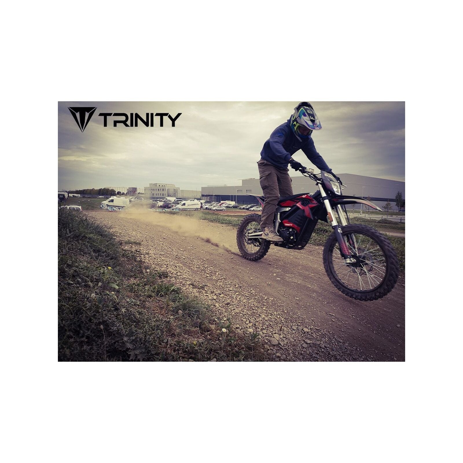Eine Person mit Helm fährt mit dem Trinity Panthera II Enduro Elektromotorrad mit Schaltgetriebe und 1143 Nm Drehmoment auf einer Schotterpiste und wirbelt dabei Staub auf. Im Hintergrund sind Gebäude und geparkte Autos zu sehen. Das Trinity-Logo erscheint in der oberen linken Ecke.