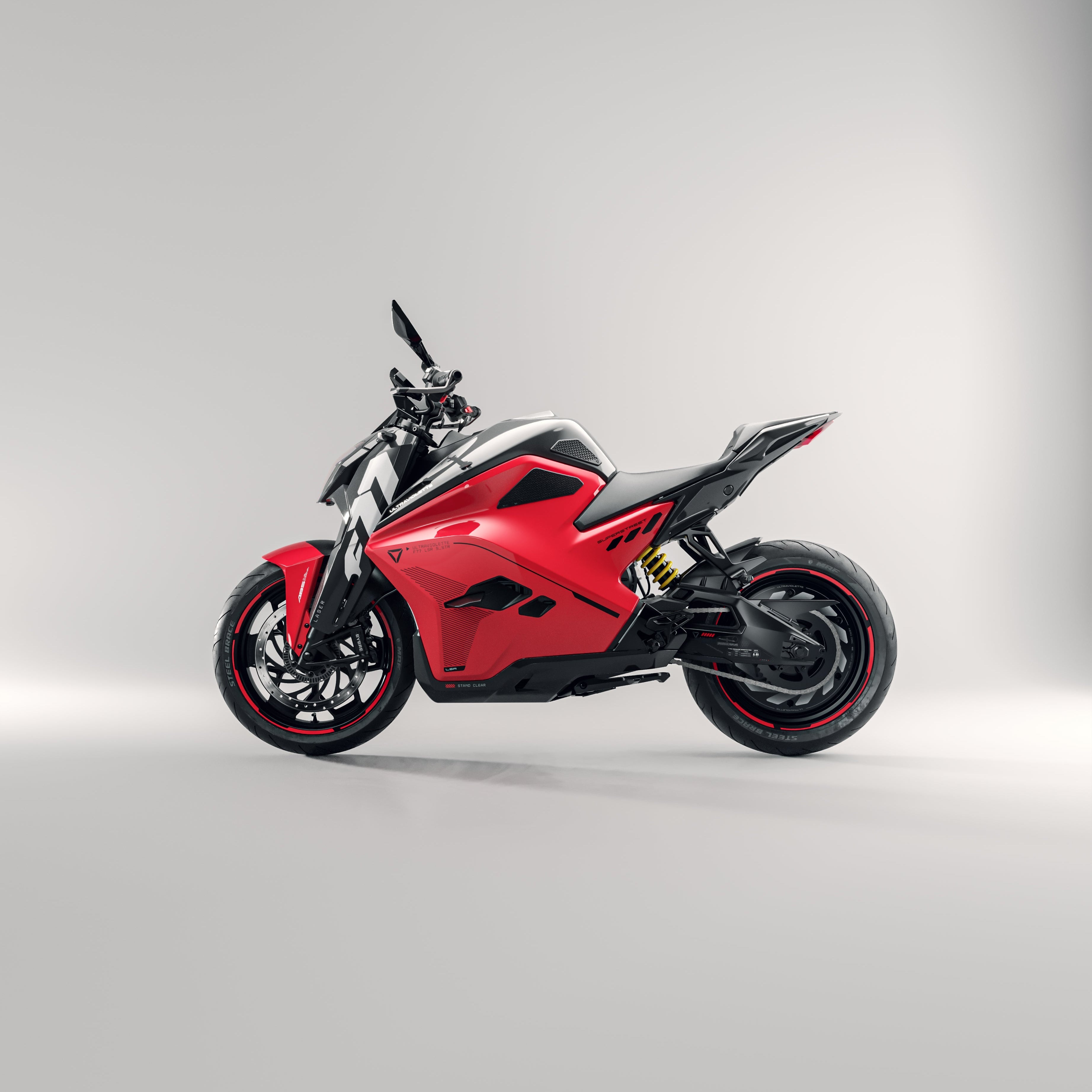 Das Ultraviolette F77 Superstreet Recon, ein sportliches rot-schwarzes Elektromotorrad von Ultraviolette, wird in einem Studio mit hellgrauem Hintergrund gezeigt. Sein modernes, aerodynamisches Design zeichnet sich durch scharfe Linien und eine sichtbare Federung in der Seitenansicht aus.