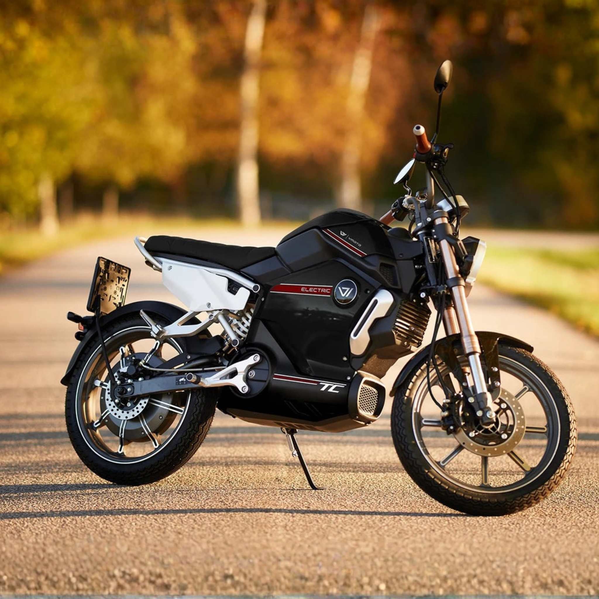 Der VMOTO TC 72V Elektroroller im Motorrad-Design von VMOTO, im schwarz-weißen Café Racer-Stil, steht auf einer sonnenbeschienenen, baumbestandenen Straße mit verschwommenem Grün und Herbstfarben im Hintergrund.