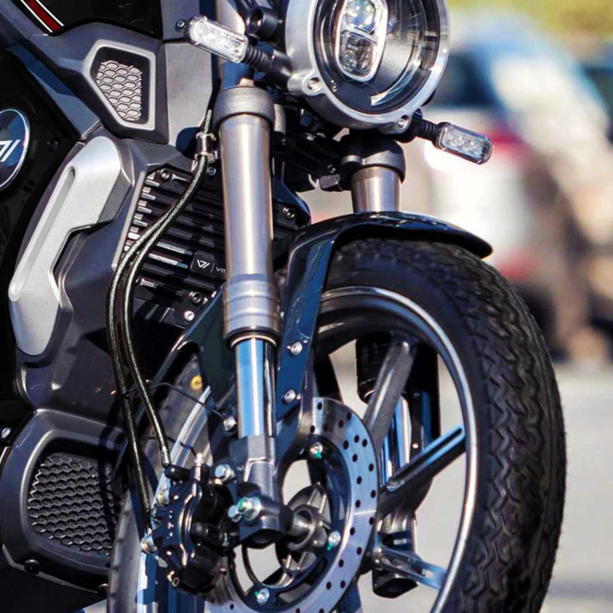 Nahaufnahme des Vorderrads, der Federgabel und des Scheinwerfers des VMOTO TC 72V Elektrorollers, die das vom Motorrad inspirierte Design zeigen. Unscharfe parkende Autos im Hintergrund verstärken das dynamische Stadtgefühl. Marke: VMOTO.