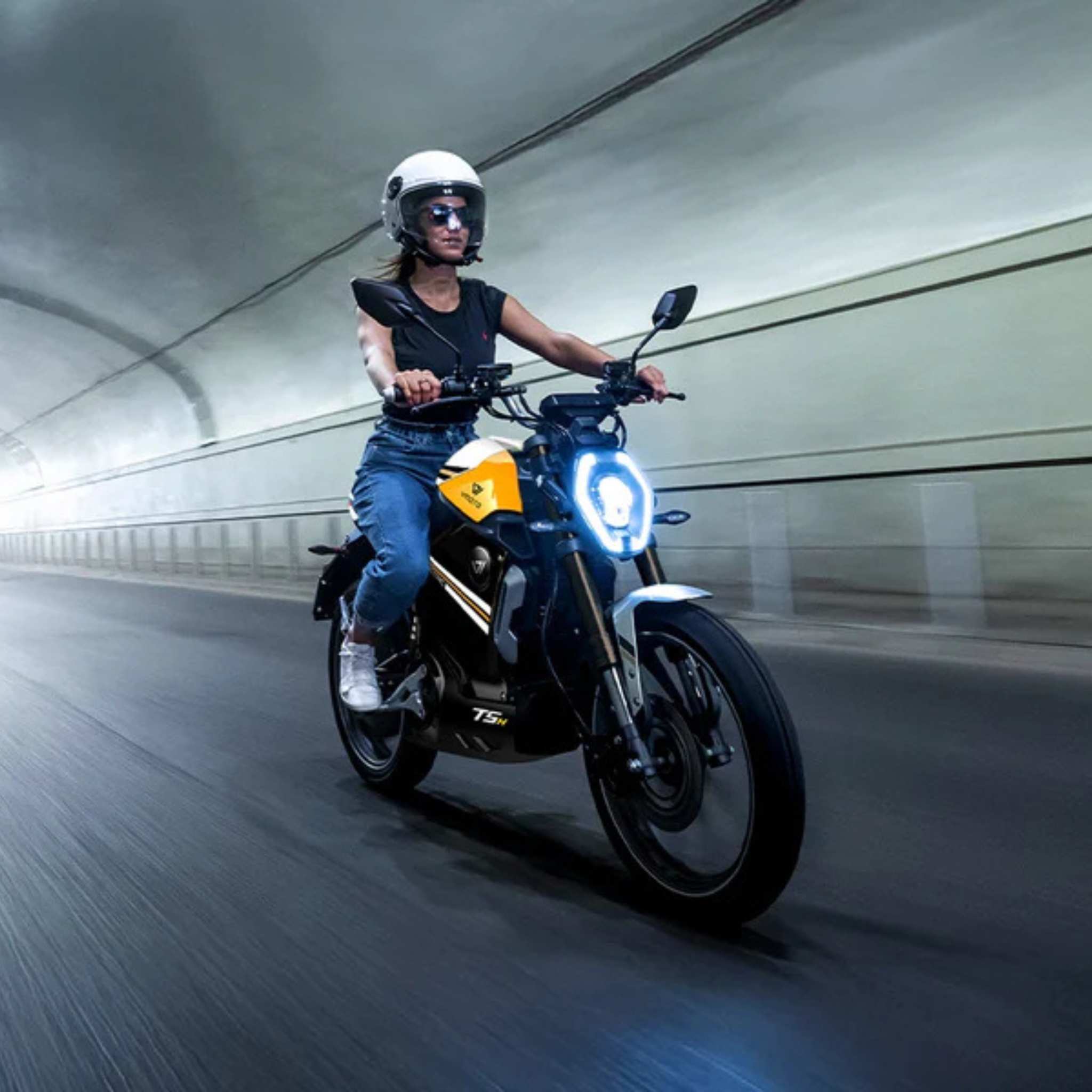 Eine Person mit weißem Helm und Sonnenbrille fährt mit dem VMOTO TSX 72V - leistungsstarker 45 km/h Elektroroller im Motorrad-Design mit hellem Scheinwerferlicht durch einen Tunnel, Bewegung verschwommen im Hintergrund.